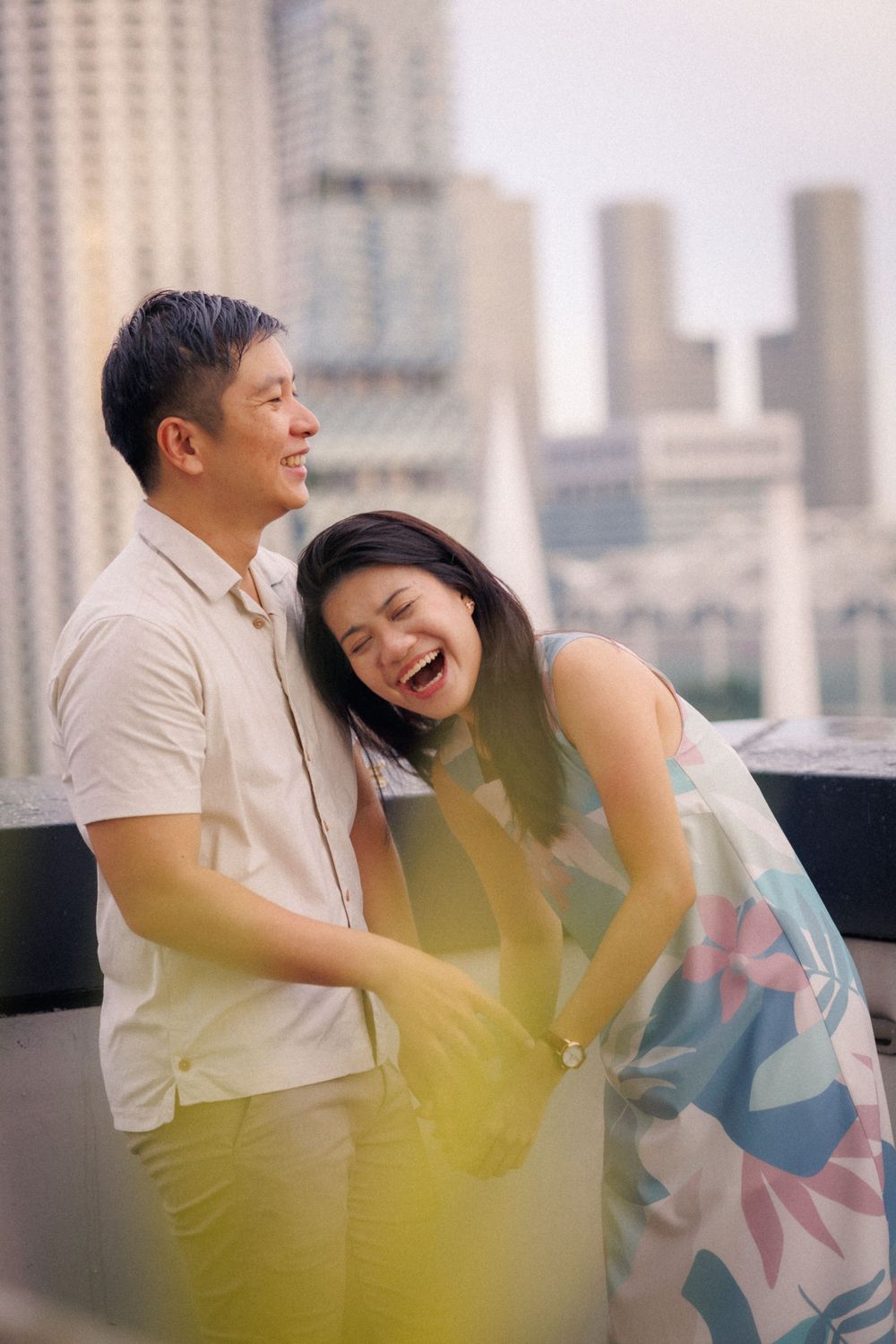 A playful couple shares a candid moment of laughter on a rooftop with a city skyline in the background.