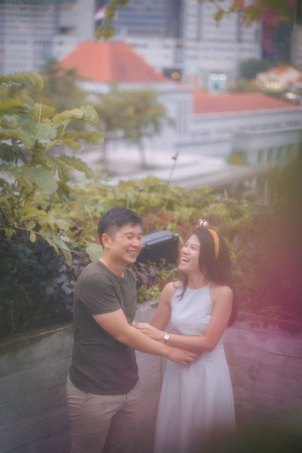 A joyful moment shared between a couple laughing together in a garden setting with urban buildings in background.