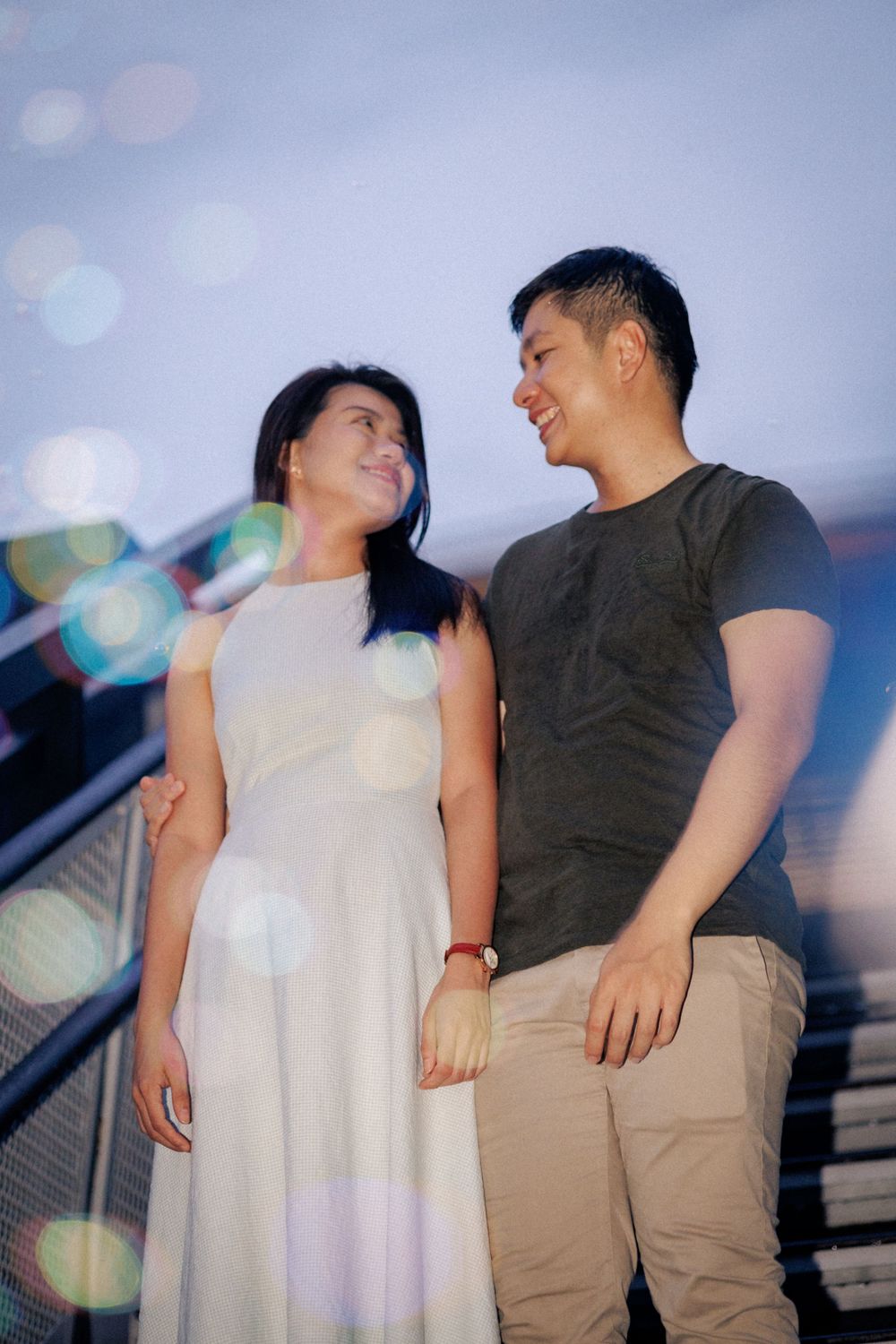 A couple shares a romantic moment outdoors at twilight with bokeh light effects in the background.