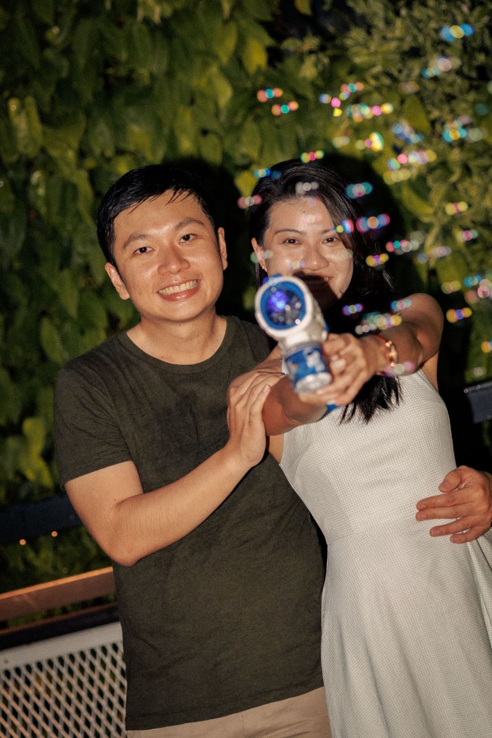 A couple plays with bubbles together during an outdoor photoshoot with greenery in the background.