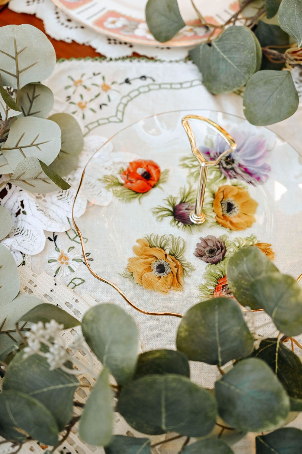 Delicate floral teacup decorated with hand-painted poppies and daisies surrounded by fresh eucalyptus leaves on lace tablecloth.