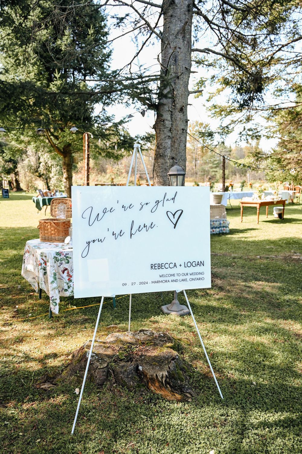 A white welcome sign on an easel at an outdoor wedding with picnic baskets and decor visible in a sunny garden setting.