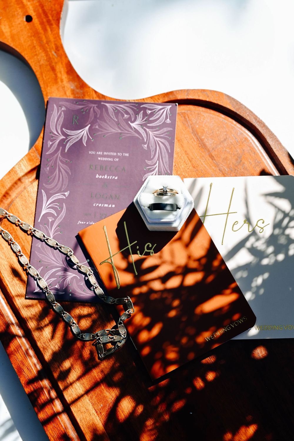 Flatlay of purple invitation card, jewelry, and perfume bottle on wooden surface with dramatic tree shadow patterns.