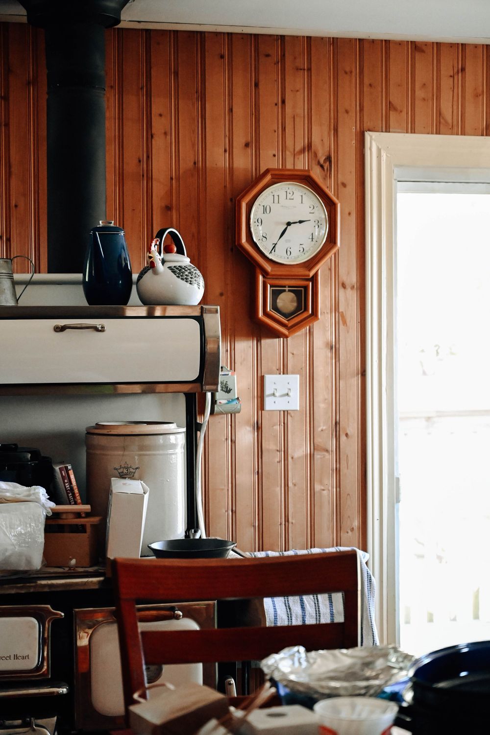 A rustic wooden wall with a vintage pendulum clock, blue ceramic pitcher, and white ceramic jug near a bright window.
