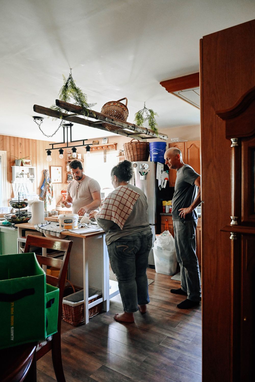 People preparing food in a rustic home kitchen with wooden walls and hanging baskets overhead.