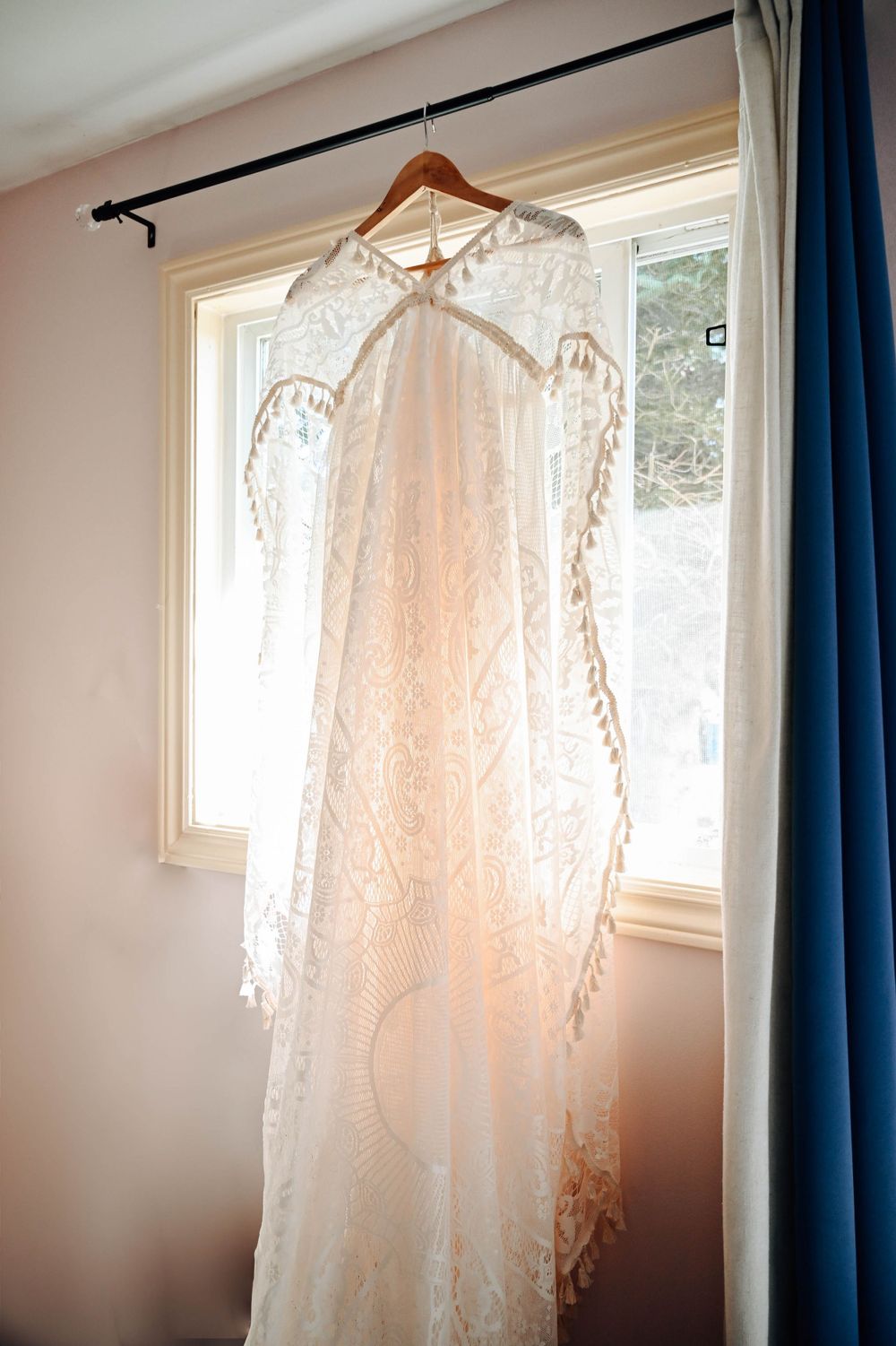 A delicate white lace wedding dress hangs in front of a sunny window with blue curtains in the background.