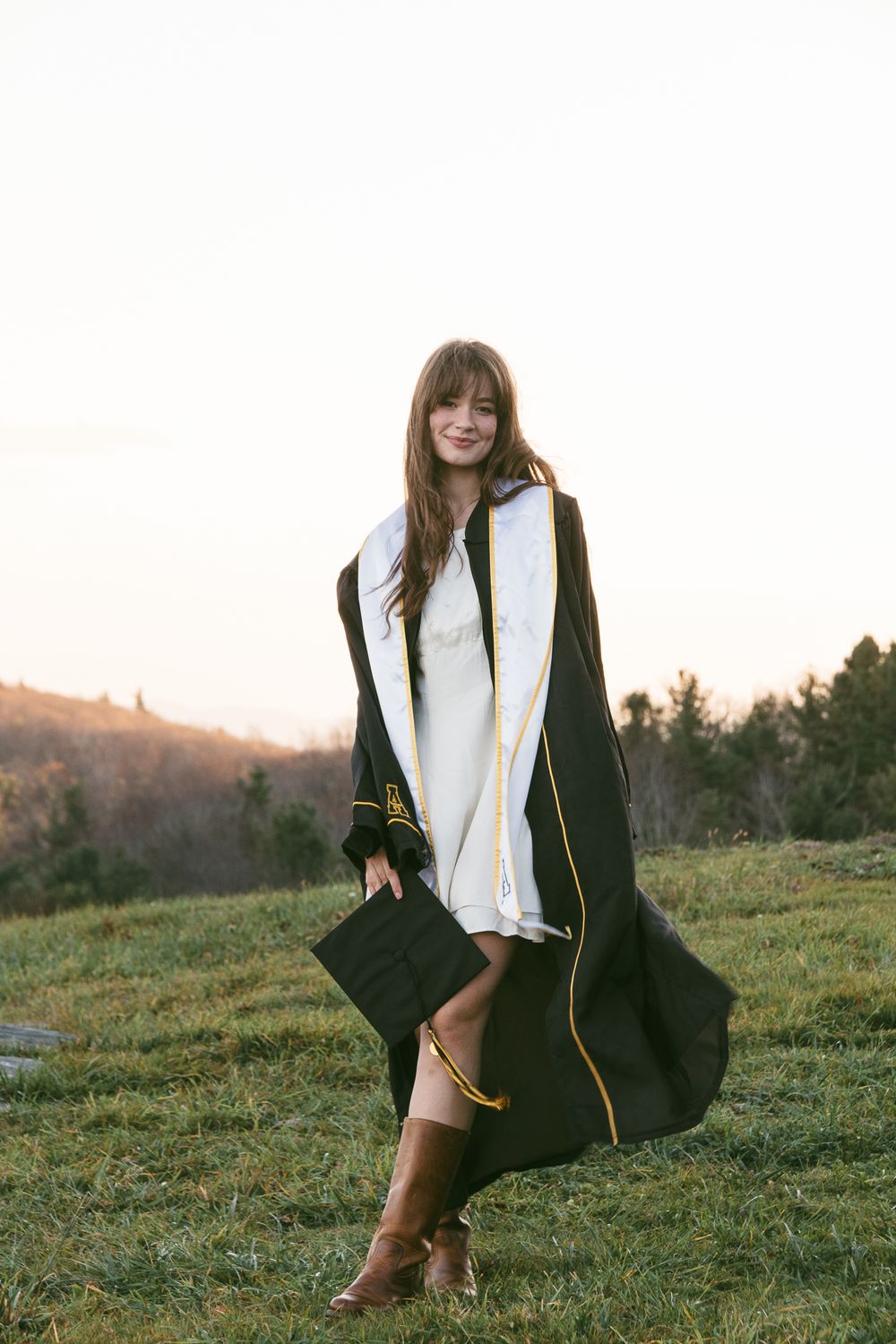 Graduate in black cap and gown poses in a grassy field at sunset. Luxury wedding photographer North Carolina