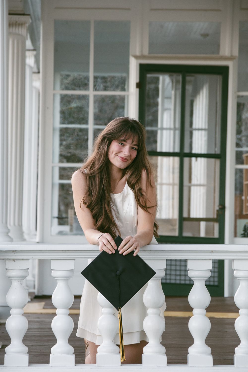 Graduate in white dress poses with graduation cap on a white porch railing. Luxury wedding photographer North Carolina