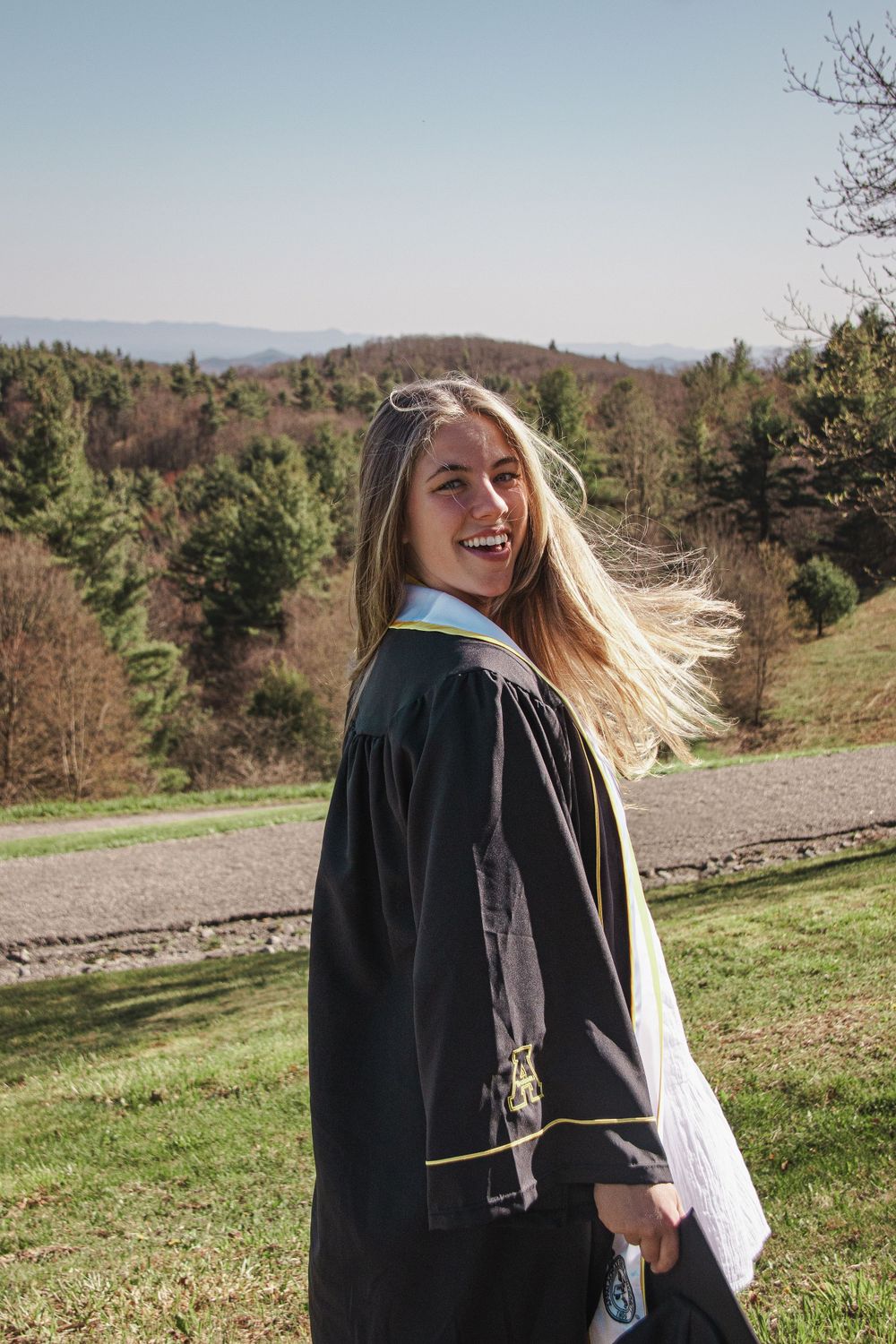 Person in graduation gown laughing with hair blowing in the wind against mountain landscape. Luxury wedding photographer North Carolina
