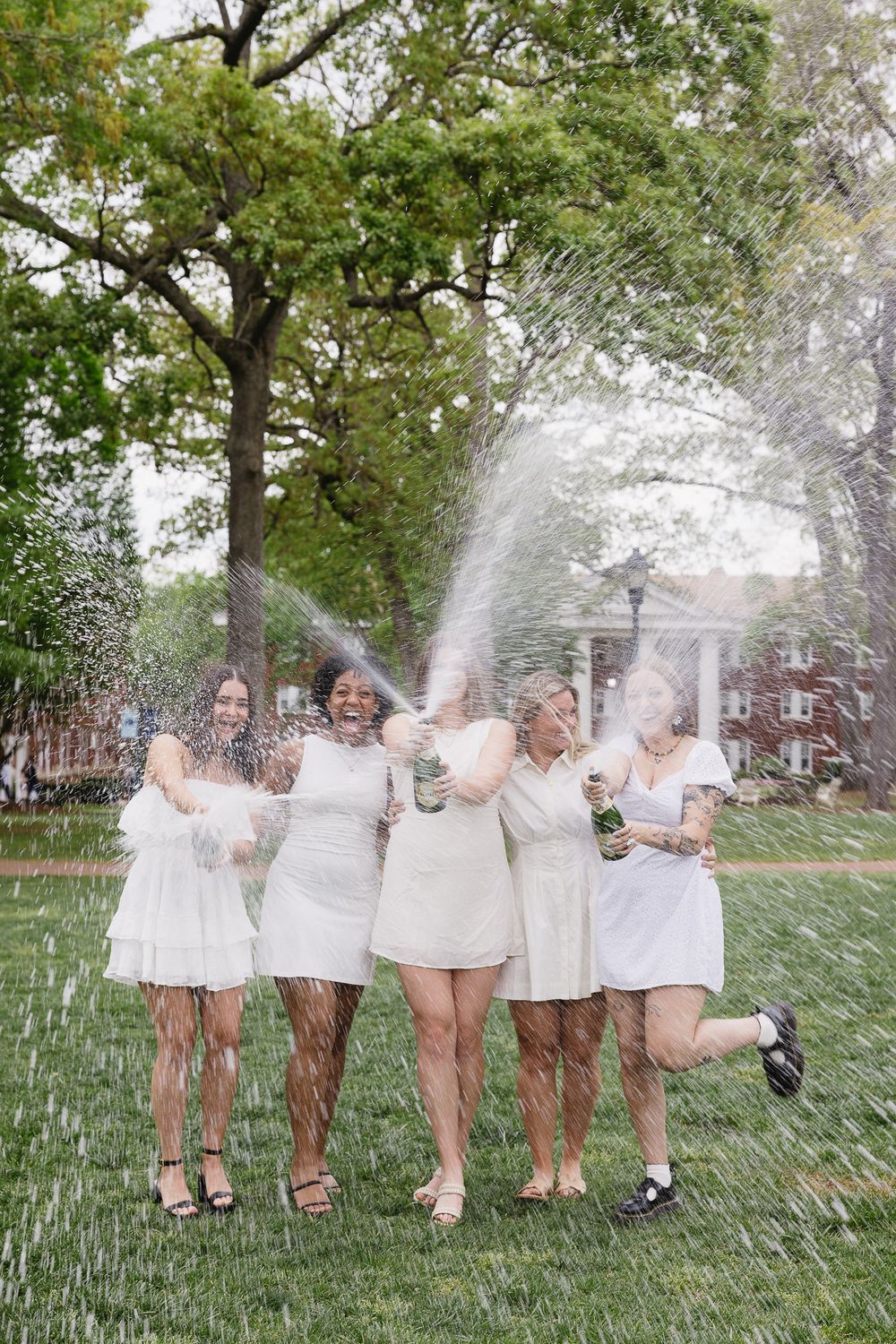 Group celebrating with champagne spray in a garden wearing white dresses. The Bradford Wedding Photographer