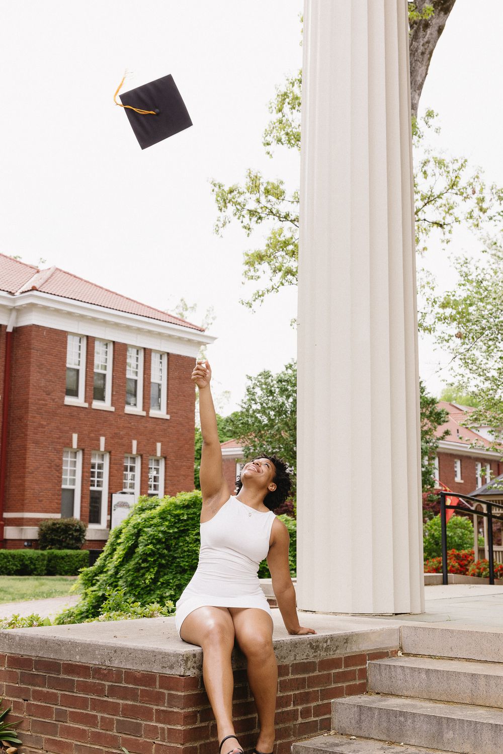 A graduate in white sits on brick steps near classical columns of a university building. The Bradford Wedding Photographer