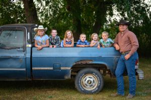 A group sits in the bed of a vintage blue pickup truck parked in a green outdoor setting.
