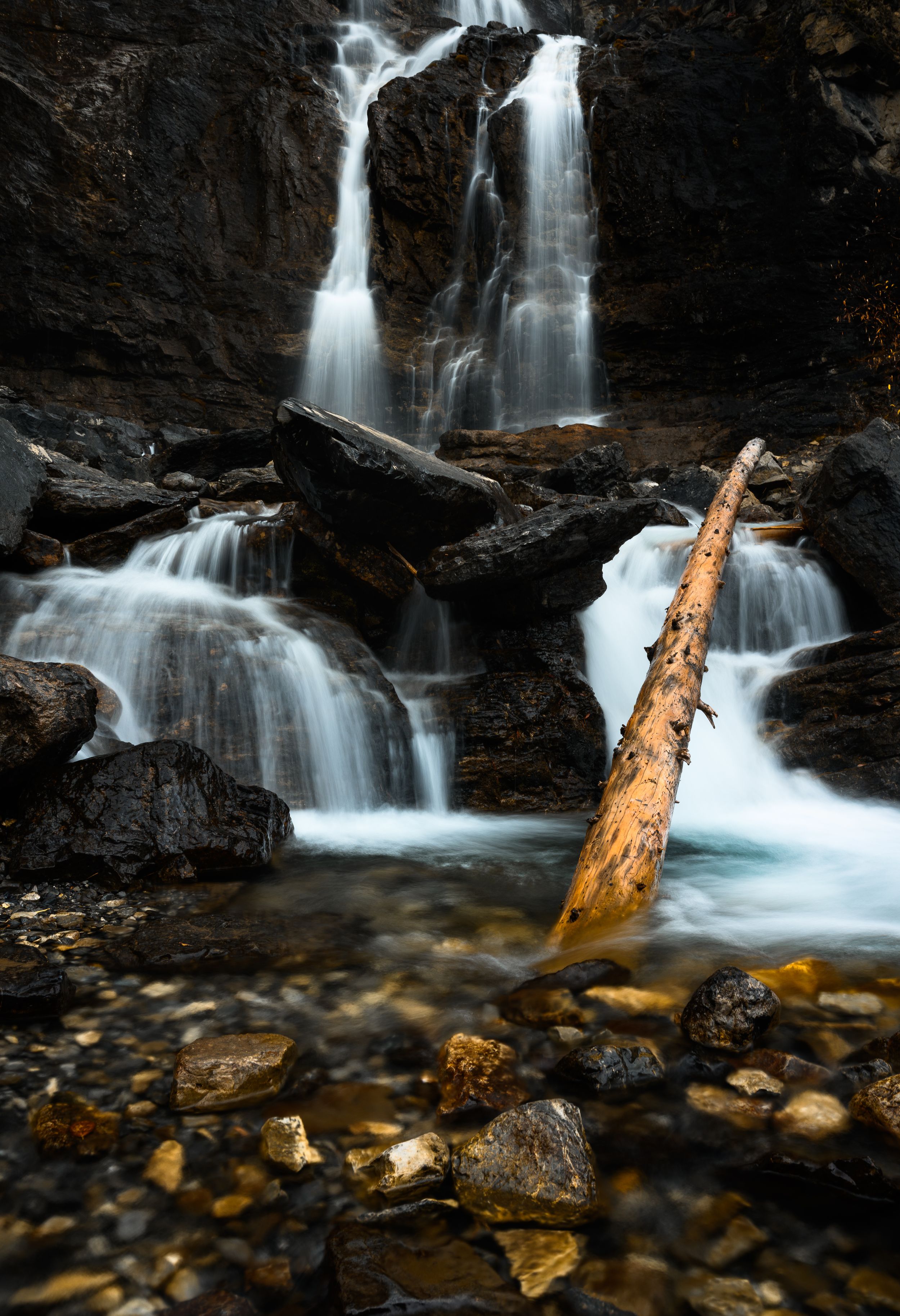 Cascading waterfall flows over dark rocky cliffs with a fallen log and river stones in the foreground.