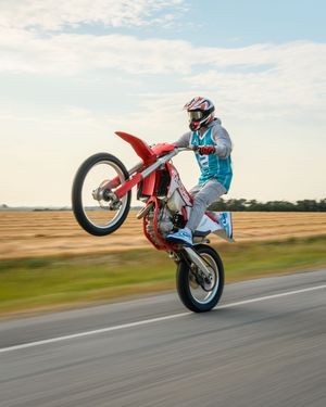 A motocross rider performs a spectacular wheelie stunt on a red dirt bike while riding along a paved road.