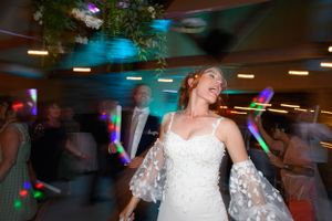 A bride in a white wedding dress dances energetically at a reception with colorful disco lights in the background.