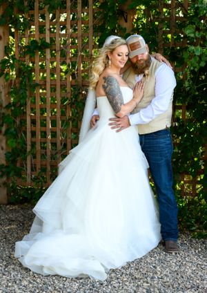 A couple embraces in front of a garden trellis, the bride wearing a flowing white gown and the groom in casual country attire.