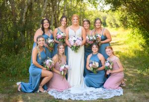 A bridal party in flowing dresses of blue and pink poses together on a sunlit wooded path.