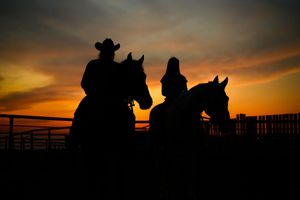 Silhouettes of cowboys on horseback against a dramatic orange sunset sky at a western ranch with fencing.