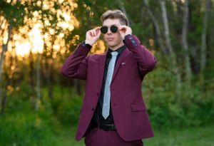 A person in a burgundy suit poses with sunglasses at sunset in a wooded area.