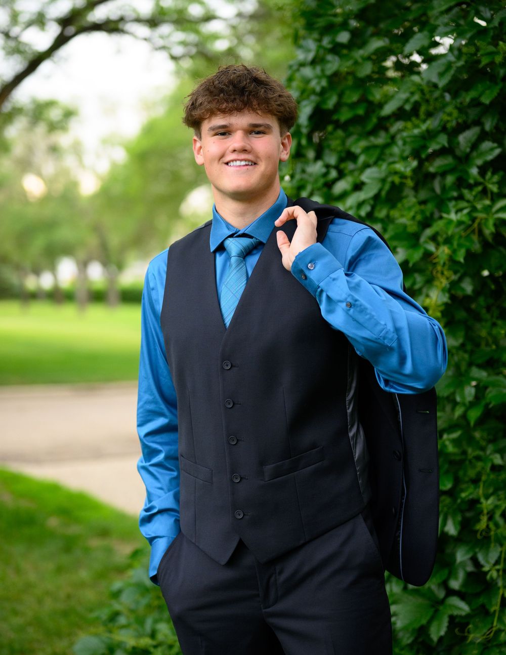 A person in a blue dress shirt and black vest poses outdoors against a green leafy background.