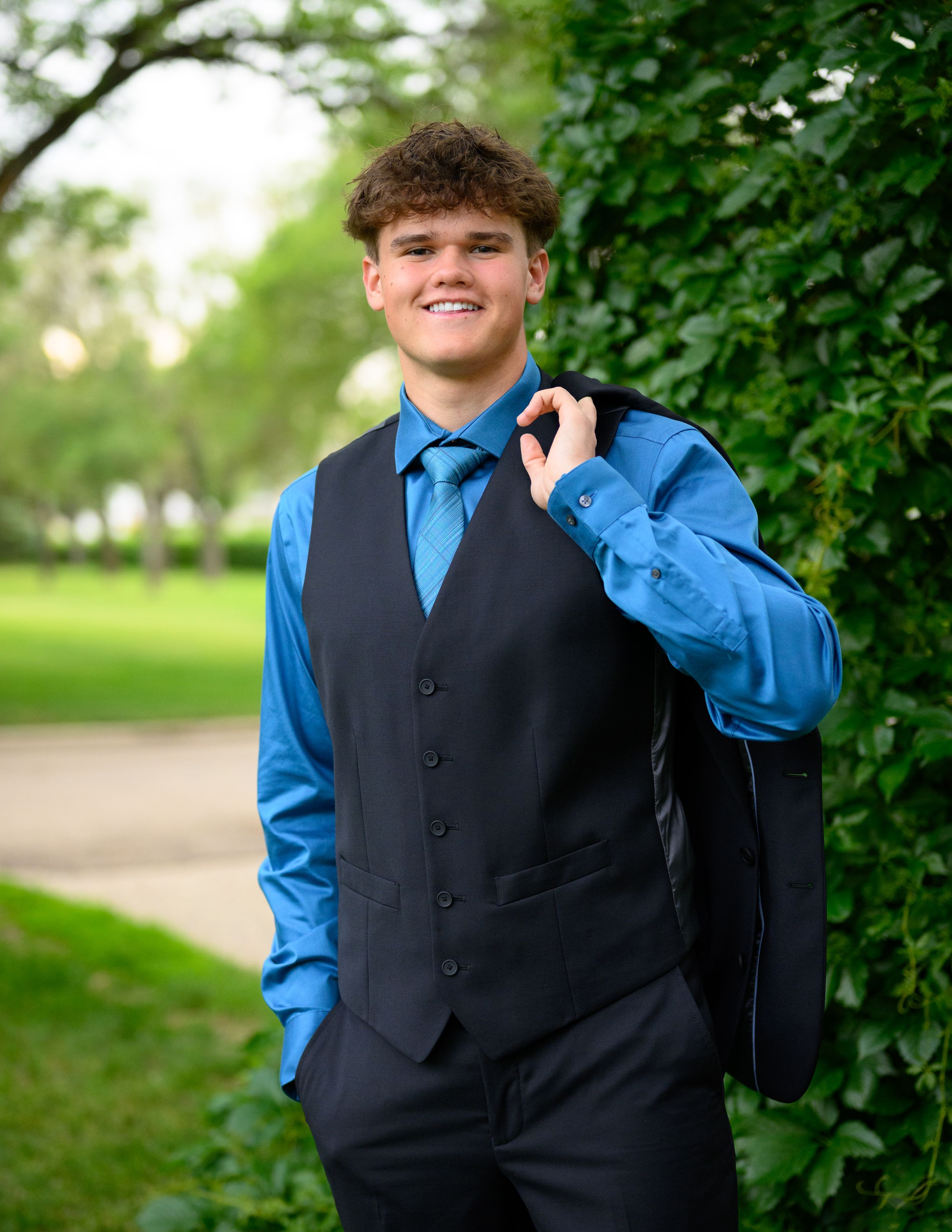 A person in a blue dress shirt and black vest poses outdoors against a green leafy background.