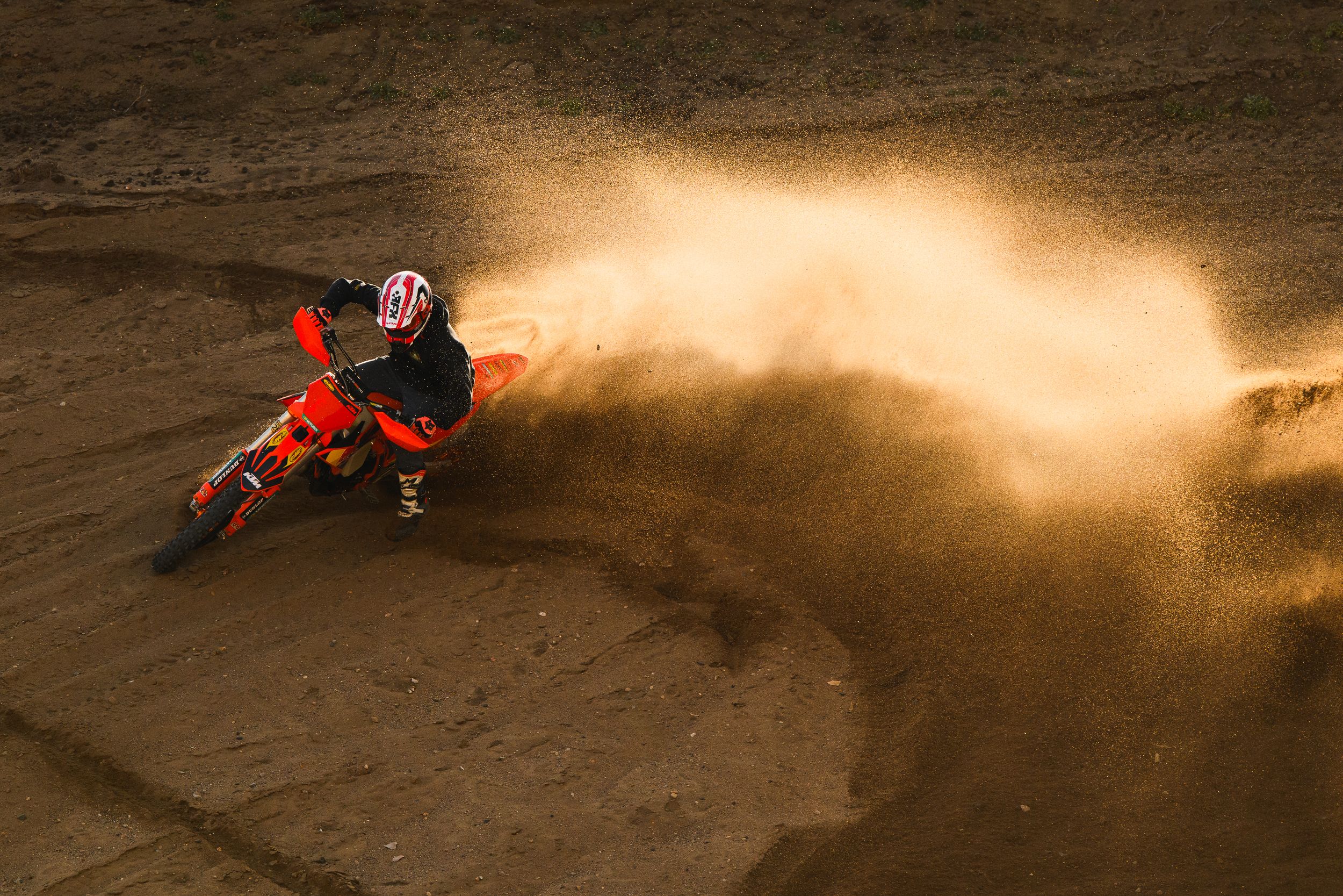 A motocross rider kicks up a dramatic cloud of dust while performing a sliding turn on a dirt track during sunset.
