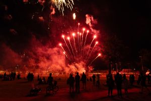 Silhouetted crowd watches bright red fireworks display illuminating night sky over beach.