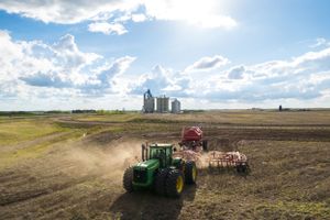 A green tractor plows a dusty field with grain silos visible in the background.