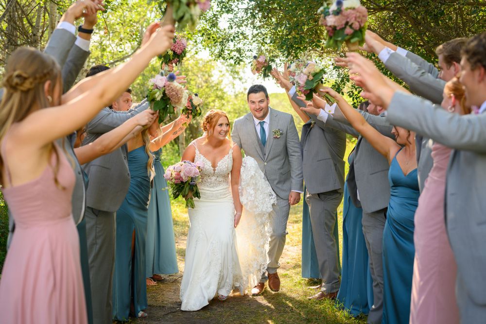 Wedding party forms an archway outdoors with hands raised holding drinks and flowers.