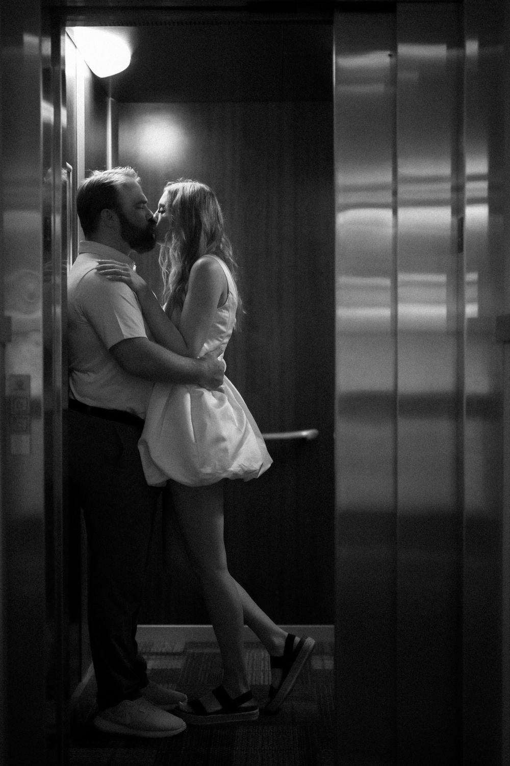 A romantic couple embraces and kisses in a dimly lit elevator, creating an intimate black and white scene.