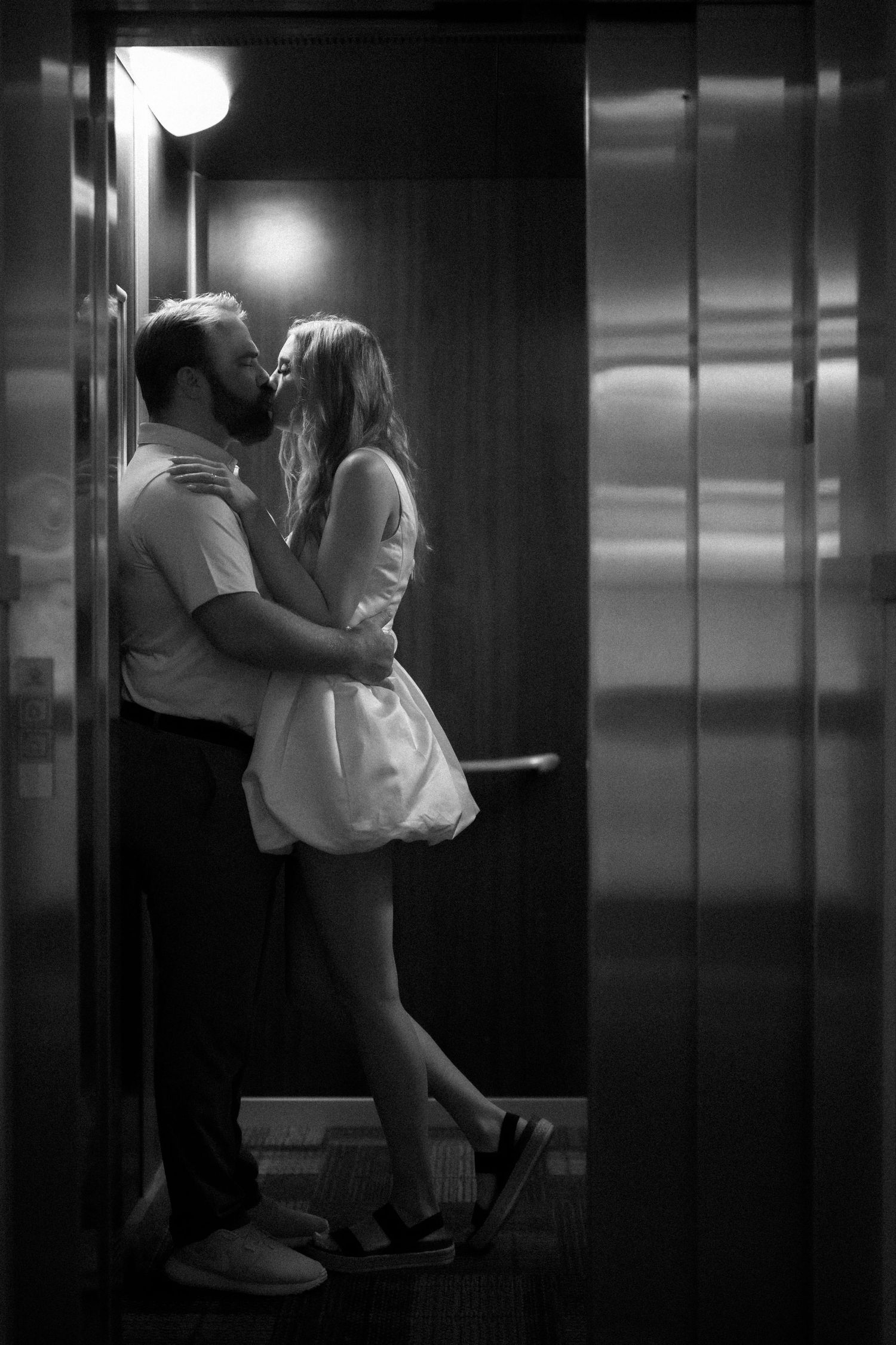 A romantic couple embraces and kisses in a dimly lit elevator, creating an intimate black and white scene.