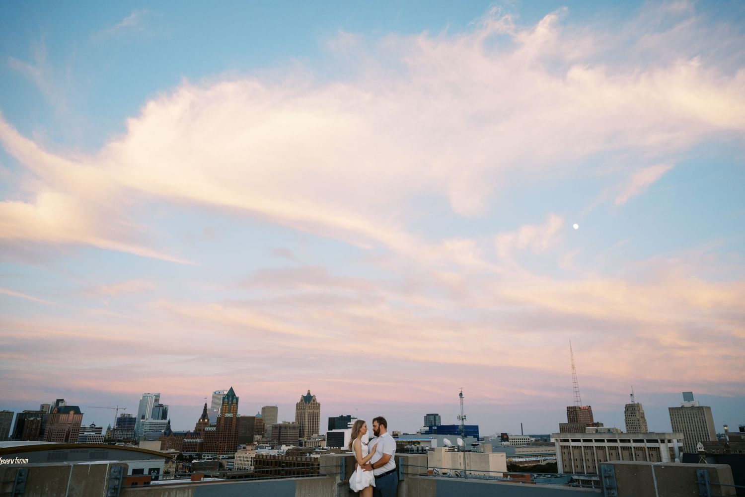 A couple embraces on a rooftop at sunset with a dramatic cityscape silhouette and pink-tinged clouds in the background.