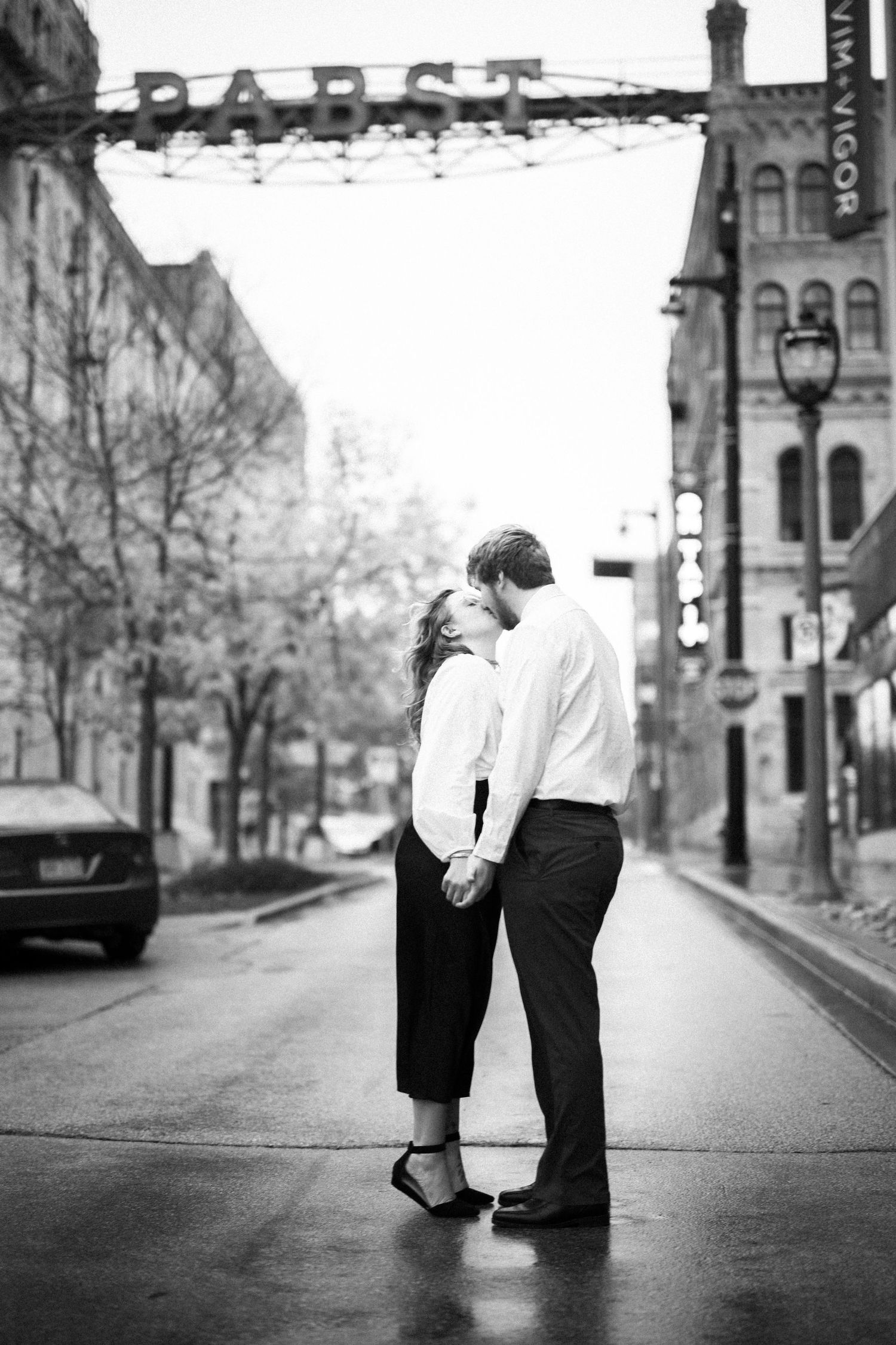 A couple embraces on a rainy city street with historic buildings and a 'Paris' sign in the background.