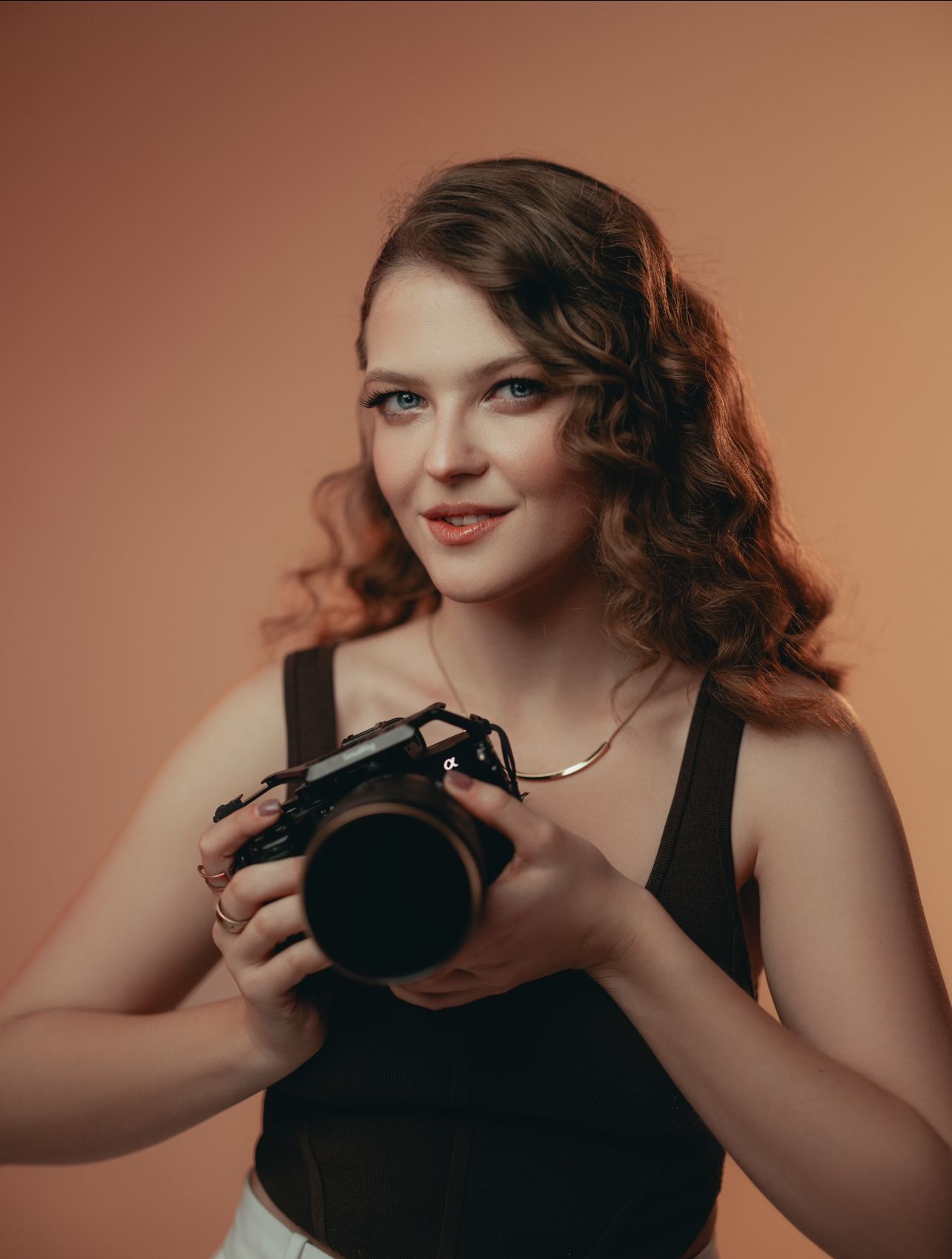 Professional photographer in black top holding camera against warm studio backdrop.