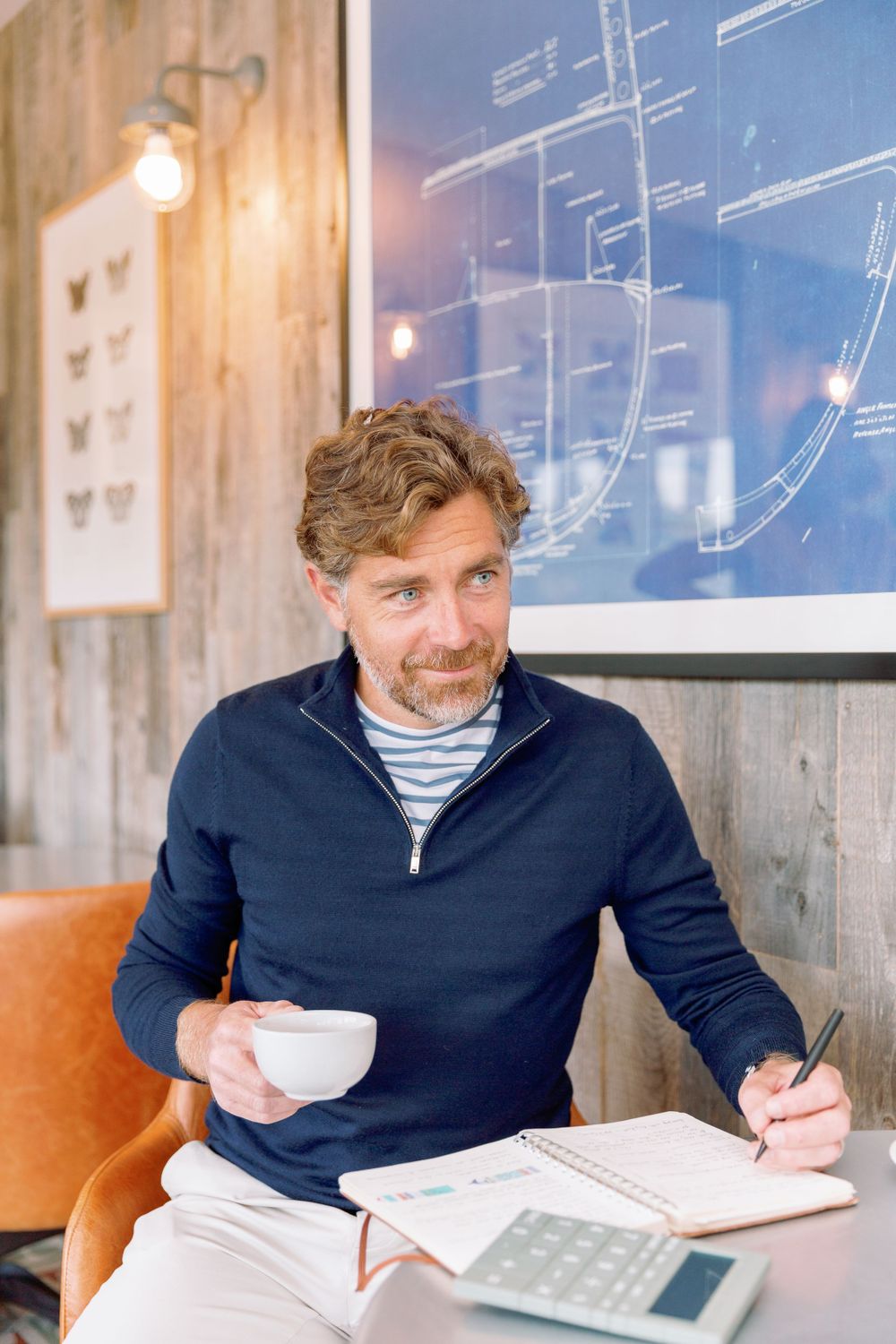 Someone in a navy sweater and striped shirt works at a table with coffee and documents in a casual cafe setting.