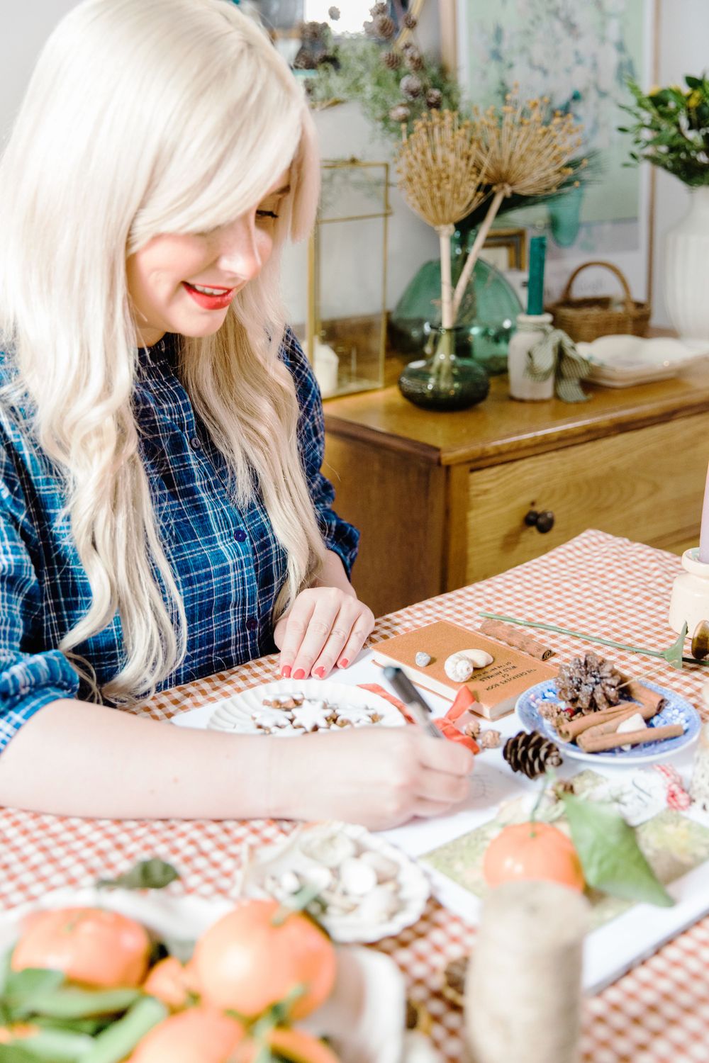 Woman with long blonde hair crafting at table with pinecones and supplies in cozy home setting.
