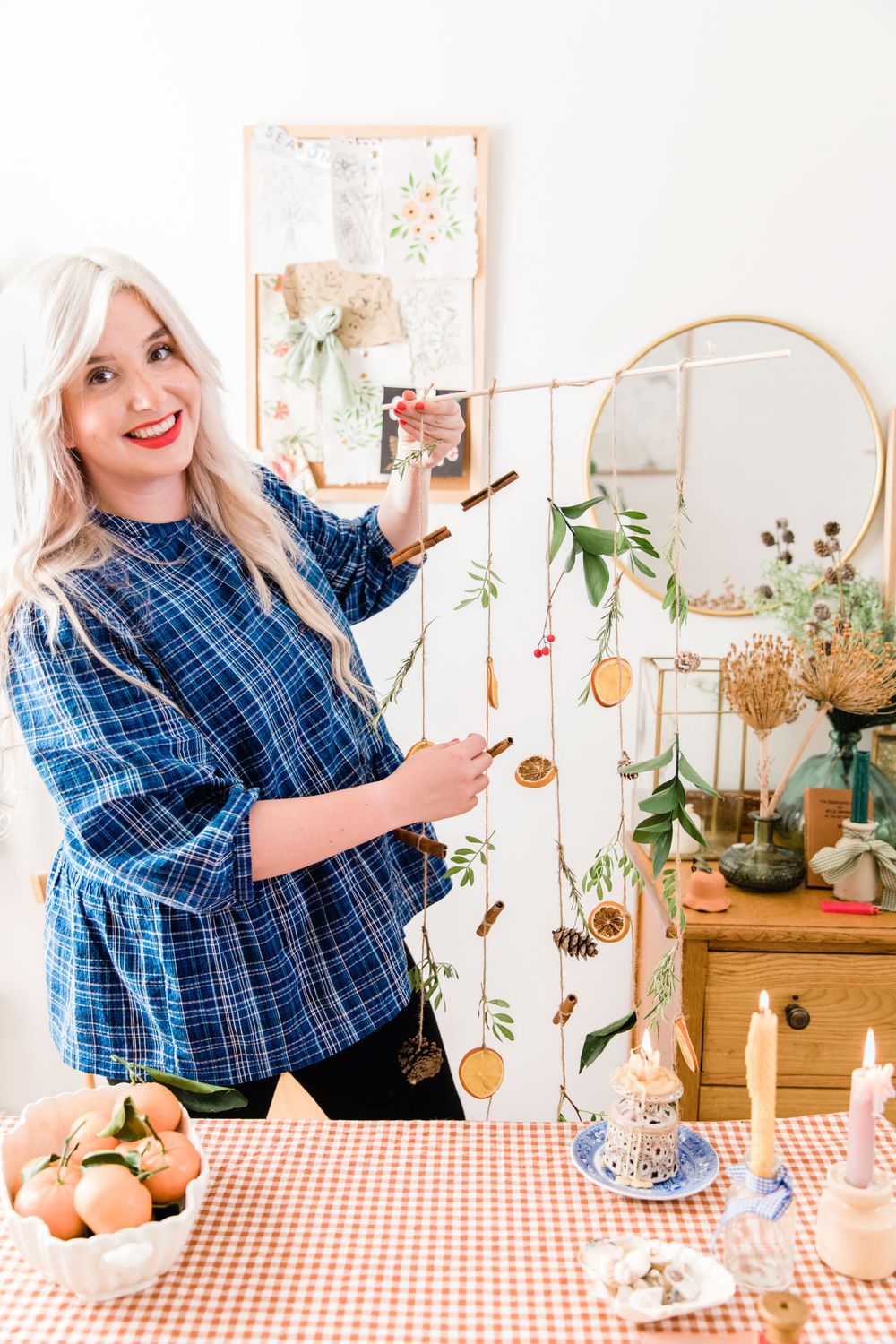Woman in blue plaid shirt decorates cozy corner with plants, frames, and natural elements on wooden table.