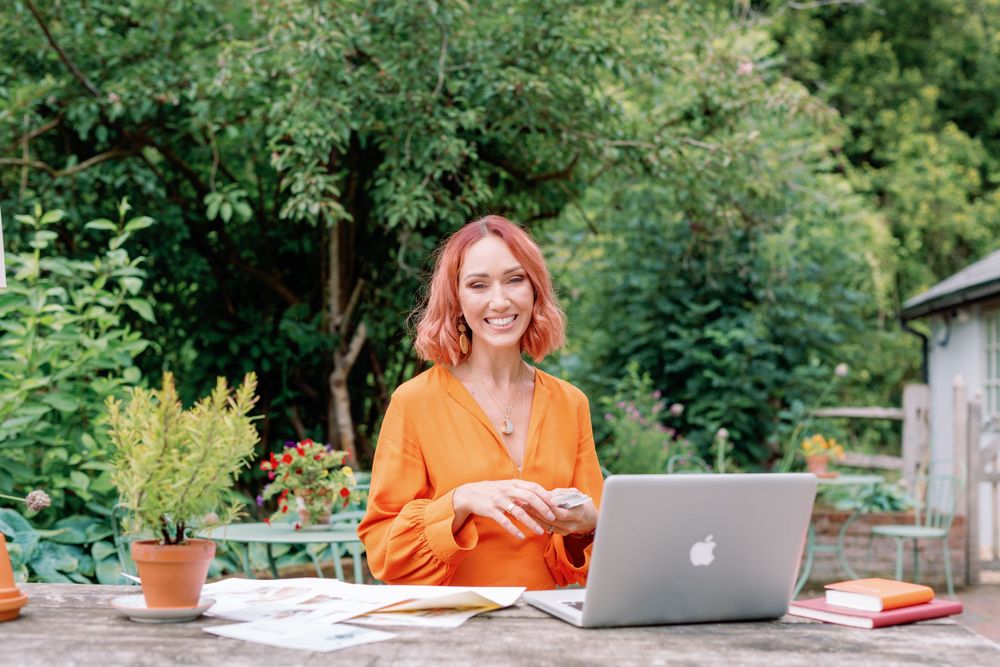 Woman in orange sweater works on laptop at outdoor garden table with potted plants.