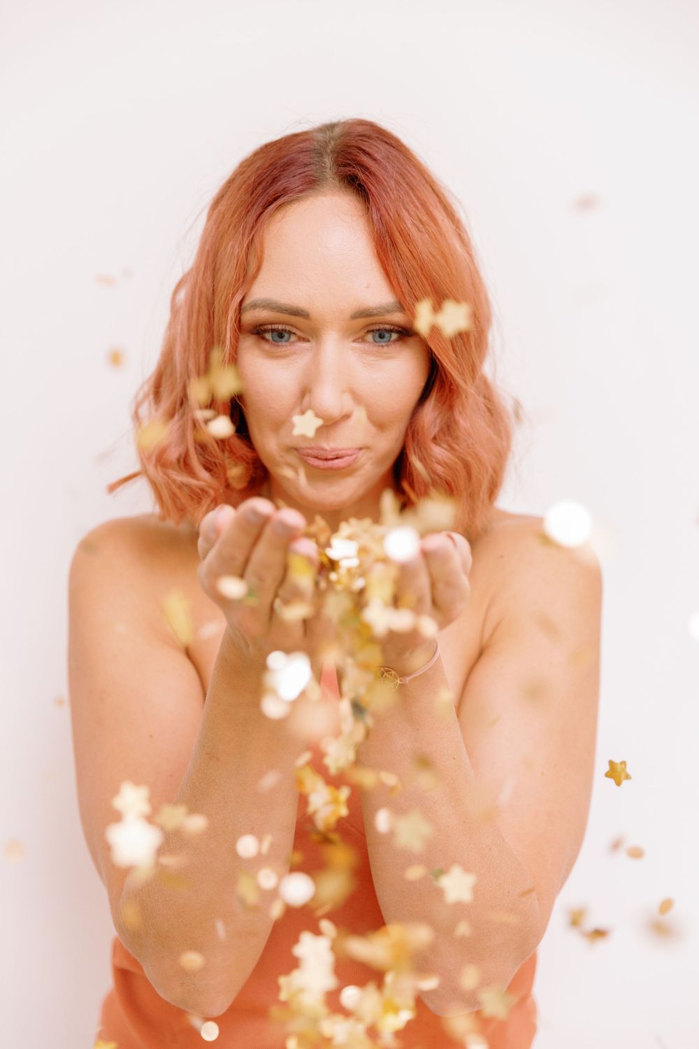 Woman blowing golden confetti stars against white background in coral-colored outfit.