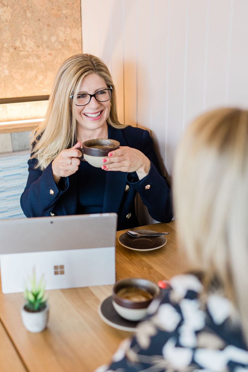 A woman with glasses smiles while holding a coffee cup at a table with a laptop and small plant.