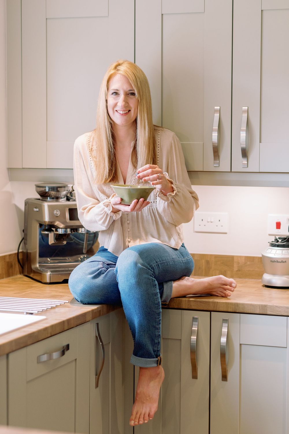 Woman in casual attire sits on kitchen counter holding drink while working from home in modern white kitchen.