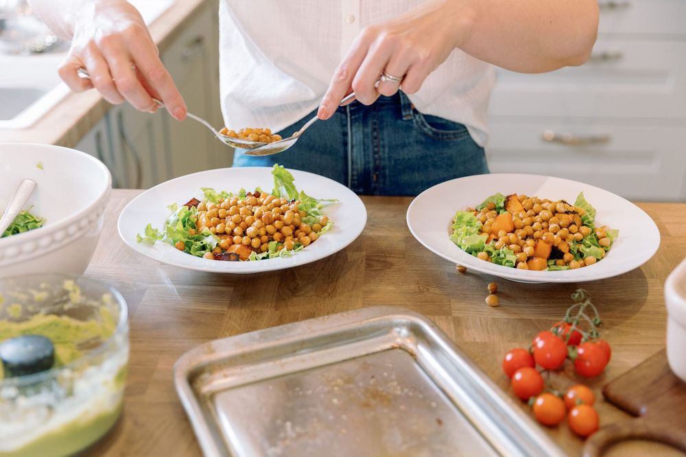 Hands seasoning a fresh salad with chickpeas and vegetables on white plates at a dining table.