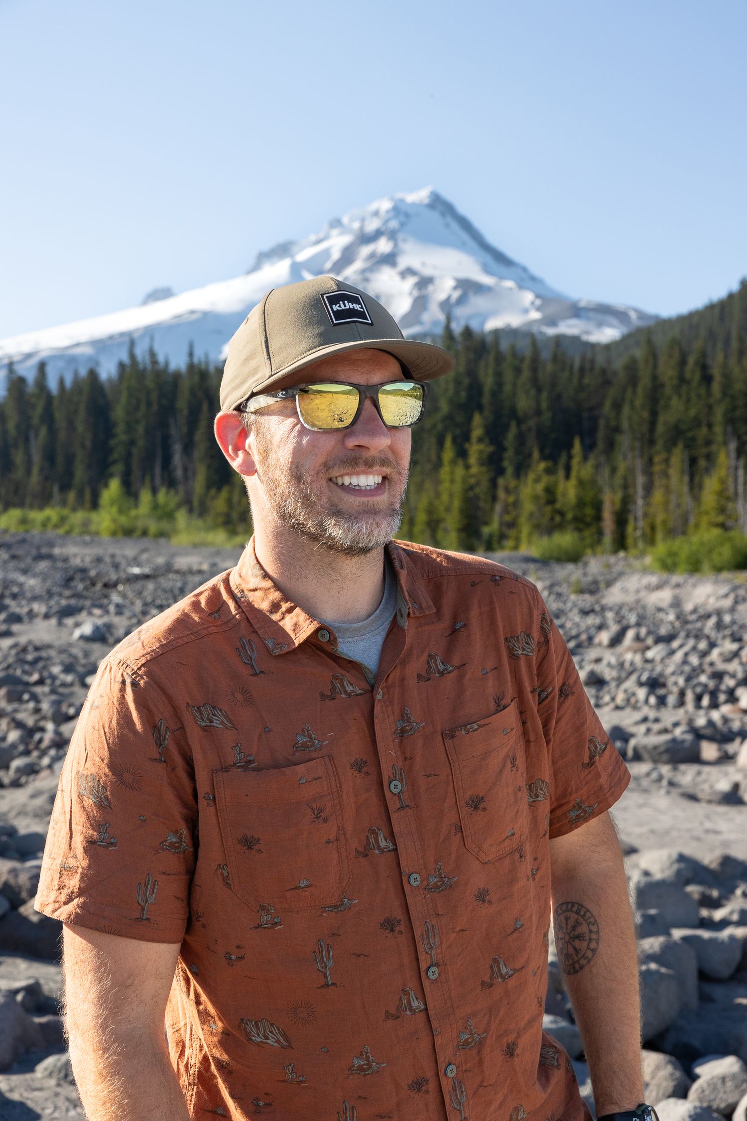 Smiling outdoor adventurer wearing sunglasses and brown patterned shirt stands in front of snow-capped Mount Hood in Oregon.