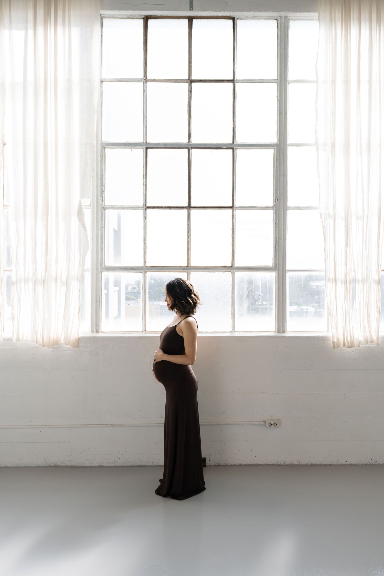 A silhouetted figure in a black dress stands contemplatively by a bright industrial window with sheer curtains.