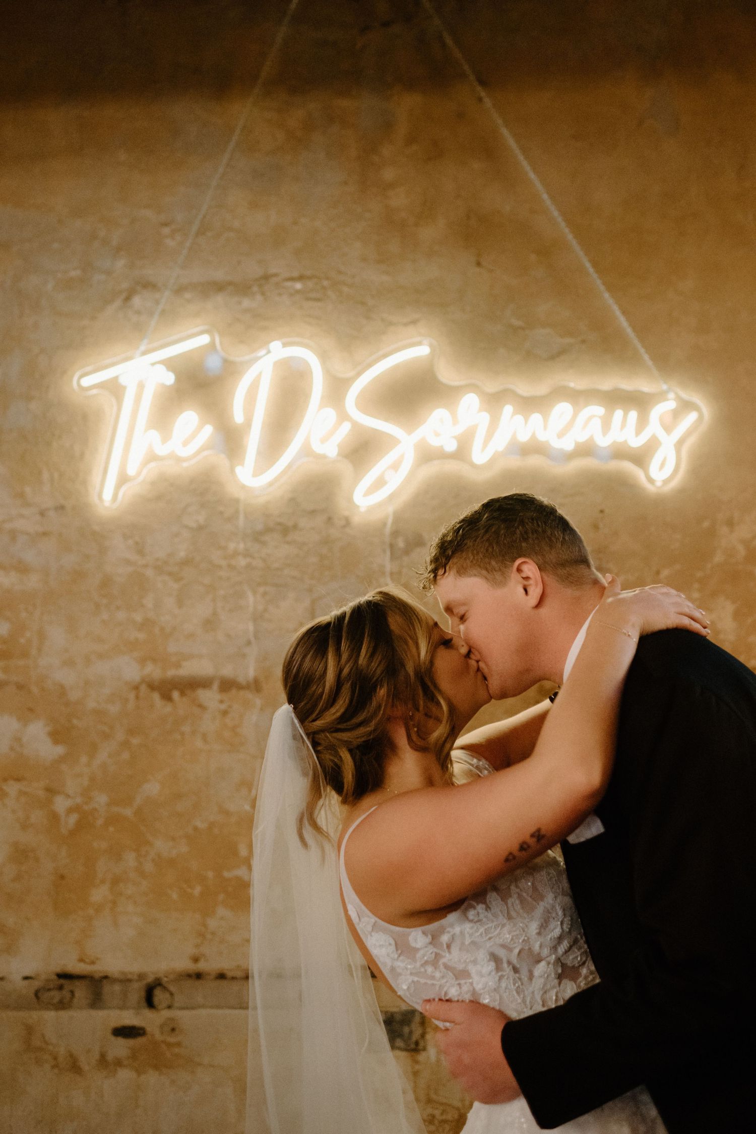 Romantic wedding kiss under glowing neon name sign against textured wall.