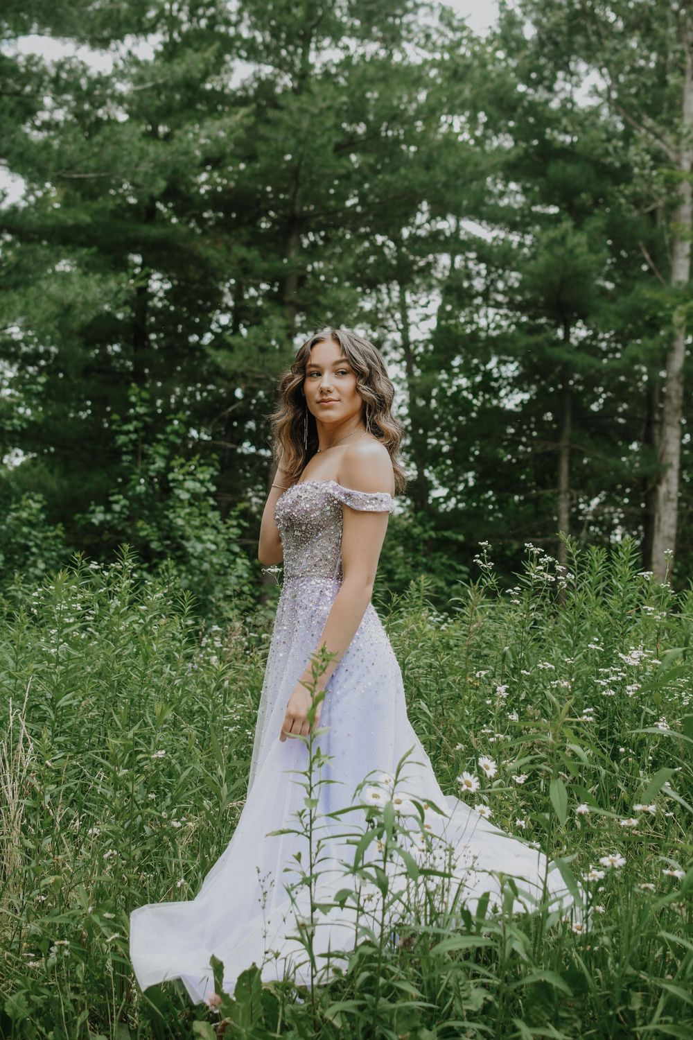 A stunning off-shoulder wedding gown shimmers among tall grass and trees in an ethereal outdoor setting.