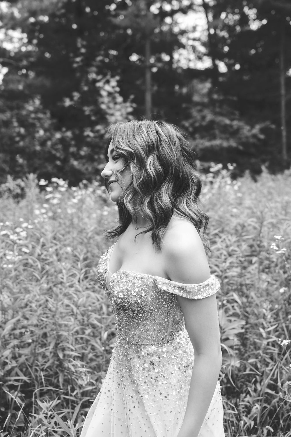 Black and white portrait of someone in a sparkly off-shoulder dress standing in a meadow with trees in the background.