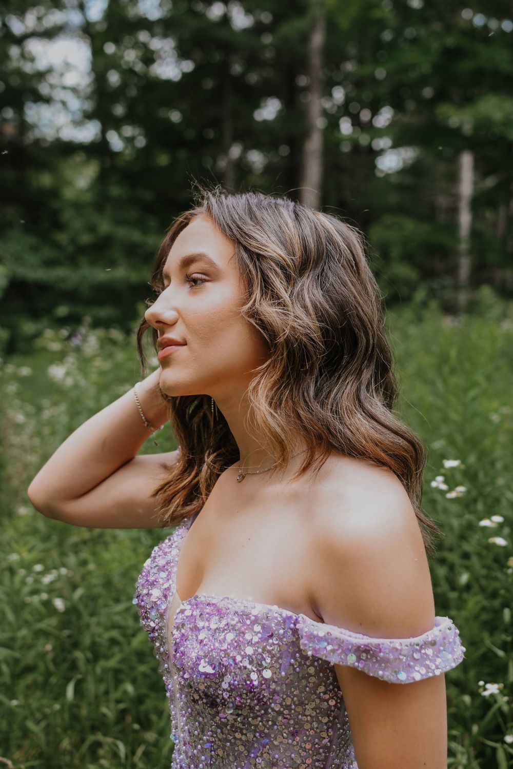 A person in a sparkling lavender off-shoulder dress poses in a lush green outdoor setting with wildflowers.