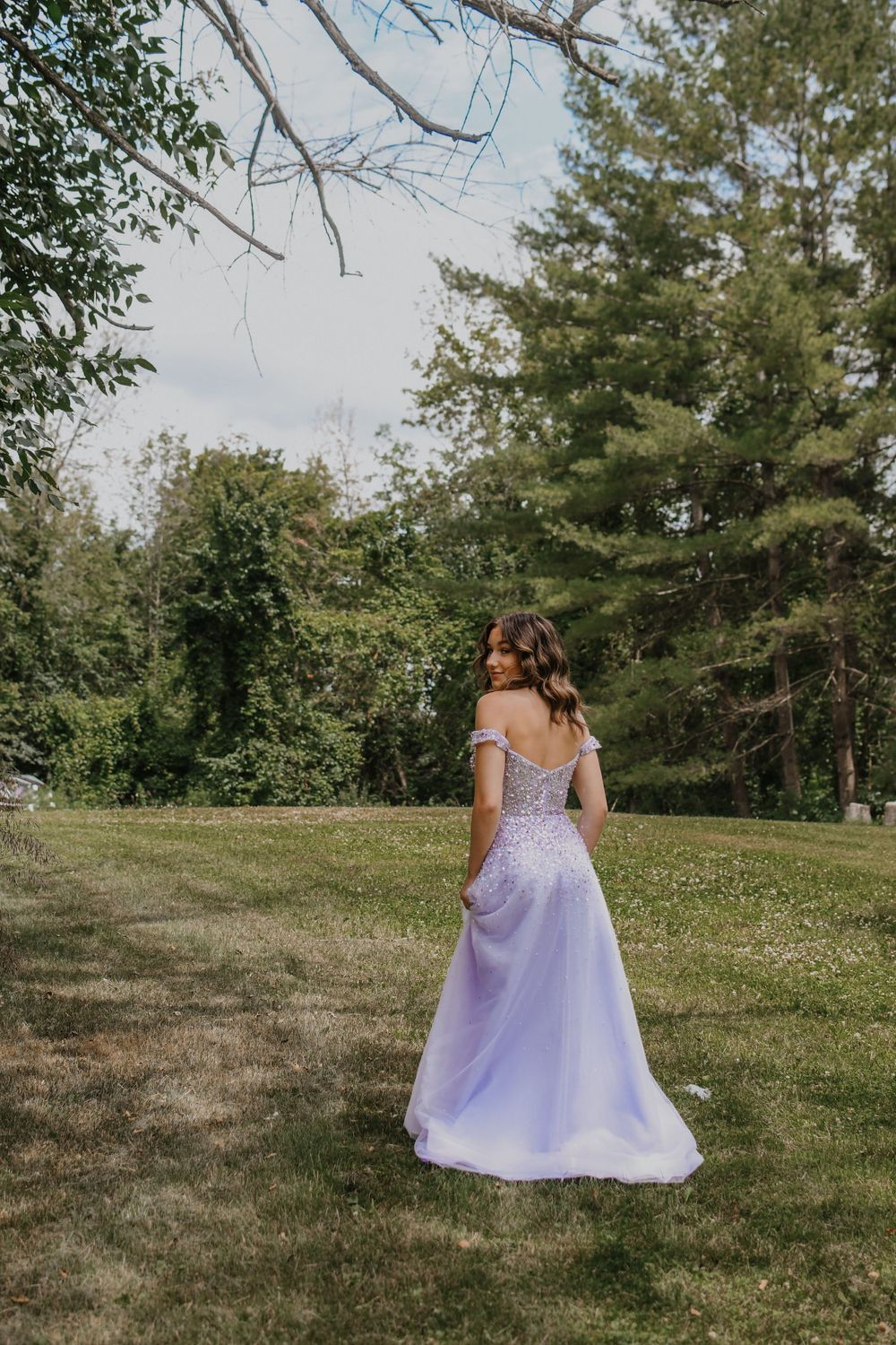 Beautiful lavender prom dress flows in a grassy outdoor setting with pine trees in the background.