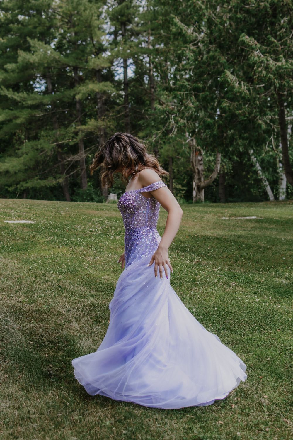Elegant off-shoulder purple gown with beaded bodice twirls gracefully on a sunny lawn surrounded by evergreens.